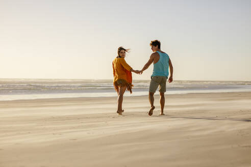 Rückansicht eines glücklichen Paares, das barfuß am Strand spazieren geht. Mann und Frau im Urlaub am Meer. - JLPSF16814