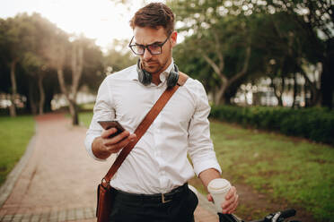 Ein Mann steht im Park und schaut auf sein Handy, während er einen Einweg-Kaffeebecher in der Hand hält. Ein Mann geht auf der Straße zum Büro, trägt eine Bürotasche und hat einen Kopfhörer um den Hals. - JLPSF16817