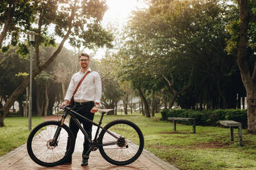 Geschäftsmann, der mit seinem Fahrrad auf einem Parkweg steht, während er ins Büro fährt. Lächelnder Mann, der mit seinem Fahrrad in einem Park steht und ins Büro fährt. - JLPSF16824