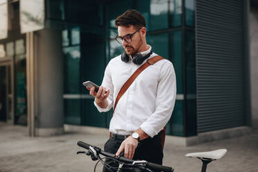 Ein Mann, der auf dem Weg ins Büro mit seinem Fahrrad auf der Straße steht und sein Mobiltelefon überprüft. Ein Geschäftsmann, der auf dem Weg ins Büro sein Mobiltelefon benutzt. - JLPSF16827