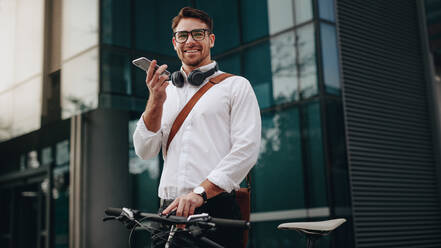 Smiling businessman talking over mobile phone standing on street with his bicycle. Man stopping by to make a call and while commuting to office on a bike. - JLPSF16829