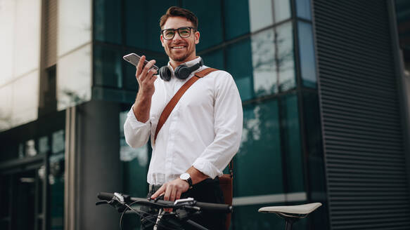 Lächelnder Geschäftsmann, der mit seinem Fahrrad auf der Straße steht und über ein Mobiltelefon spricht. Ein Mann hält an, um einen Anruf zu tätigen, während er mit dem Fahrrad ins Büro fährt. - JLPSF16829
