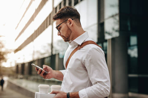 Seitenansicht eines Geschäftsmannes, der ein Mobiltelefon benutzt und Kaffeetassen auf der Straße hält. Ein Mann steht vor einem Gebäude und hält zwei Kaffeetassen, während er sein Mobiltelefon überprüft. - JLPSF16830