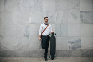 Formell gekleideter Mann auf der Straße mit einem Longboard. Geschäftsmann, der im Freien an einer Wand steht und ein langes Skateboard hält. - JLPSF16834