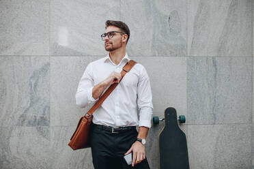 Businessman wearing an office bag standing against a wall holding mobile phone. Formally dressed man standing with a long skateboard looking away. - JLPSF16835