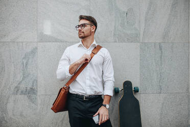 Geschäftsmann mit Bürotasche, der an einer Wand steht und ein Handy in der Hand hält. Ein formell gekleideter Mann steht mit einem langen Skateboard und schaut weg. - JLPSF16835