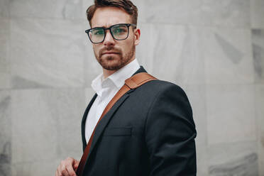 Close up portrait of young businessman standing beside a wall. Side view of a man wearing eyeglasses and carrying an office bag standing outdoors. - JLPSF16836