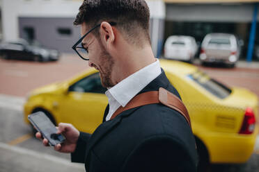 Side view of a businessman walking on street looking at his mobile phone. Smiling man in formal clothes standing on street using mobile phone with a cab in the background. - JLPSF16837