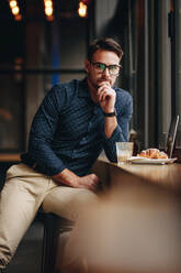 Freiberuflicher Geschäftsmann in einem Restaurant mit Essen und Laptop vor sich. Mann sitzt in einem Café mit Snacks und Laptop und denkt nach. - JLPSF16854