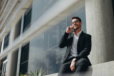 Businessman taking a break from work talking on mobile phone sitting outdoors. Businessman in formal clothes sitting outside a building and talking on mobile phone. - JLPSF16866