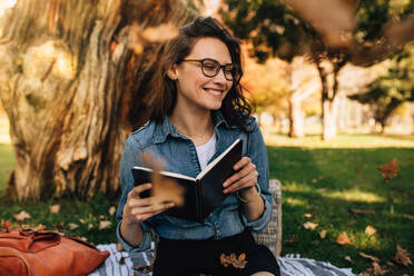 Lächelnde Frau, die im Park sitzt und ein Buch in der Hand hält, während Blätter um sie herum fallen. Schöne Frau mit Brille, die wegschaut und lächelt, mit einem Buch in der Hand. - JLPSF16869