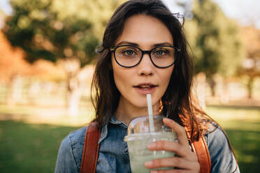 Nahaufnahme einer jungen Frau im Park, die einen Saft trinkt. Eine Studentin mit Brille trinkt einen Smoothie und schaut in die Kamera. - JLPSF16870