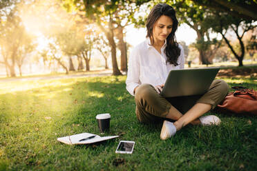 Junge Geschäftsfrau, die im Park an ihrem Notebook arbeitet. Freiberuflerin, die auf einer Wiese sitzt und einen Laptop benutzt, mit ihrem Tagebuch, ihrem Handy und einem Kaffee an ihrer Seite auf der Wiese. - JLPSF16872