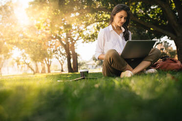 Frau sitzt im Park im Gras und arbeitet an einem Laptop. Frau mit Kopfhörern, die einen Laptop benutzt, während sie unter einem Baum im Park sitzt und das helle Sonnenlicht von hinten sieht. - JLPSF16873