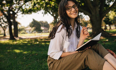Schöne junge Frau mit Brille sitzt im Park und schreibt in ein Buch. Kaukasische Frau schreibt in einem Buch im Park. - JLPSF16874