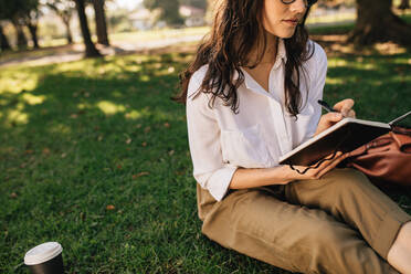 Frau sitzt mit Buch im Gras im Park und macht sich Notizen. Frau entspannt sich im Freien im Park und schreibt in ein Tagebuch. - JLPSF16875