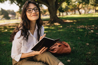 Hübsche Frau schreibt im Park in ein Buch. Frau mit Brille sitzt im Park mit einem Buch. - JLPSF16876