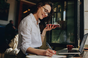 Lächelnde Frau, die Notizen macht und telefoniert, während sie in einem Café sitzt. Freiberufler, der in einem Café arbeitet. - JLPSF16882