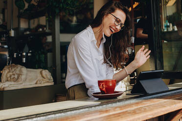 Woman on video call using a digital tablet sitting in cafe. Smiling businesswoman at coffee shop making gestures on a video call. - JLPSF16884