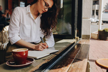 Frau sitzt am Fenster eines Cafés und schreibt in ein Buch, daneben steht eine Tasse Kaffee. Frau mit weißem Hemd und Brille sitzt im Café und macht sich Notizen. - JLPSF16886