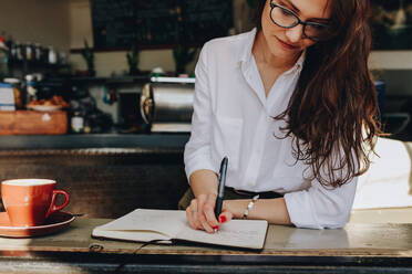 Frau im Café schreibt Notizen in ein Buch, während sie im Café sitzt. Frau macht einige Notizen in ihrem Tagebuch im Café. - JLPSF16888