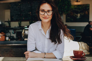 Porträt einer attraktiven Frau, die in einem Café sitzt, mit einem Notizbuch und einer Tasse Kaffee auf dem Tisch. - JLPSF16889