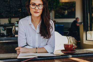 Porträt einer selbstbewussten jungen Frau, die mit einem Buch und einer Tasse Kaffee auf dem Tisch in einem Café sitzt. Kaukasische Frau entspannt sich im Café. - JLPSF16890