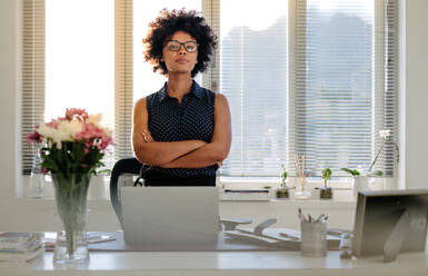 Portrait of confident young businesswoman standing at her desk with arms crossed and looking at camera. African woman with curly hair standing at her workplace. - JLPSF16899