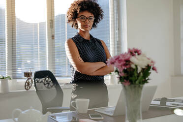 Casually dressed african business woman looking into the camera with her arms crossed while standing in front of her desk. Female with curly hair wearing eyeglasses at her workplace. - JLPSF16900