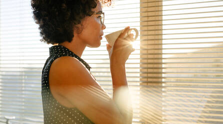 Junge afrikanische Geschäftsfrau trinkt Kaffee, während sie an einem Fenster im Büro steht. Frau schaut nach draußen und denkt über etwas nach, während sie Kaffee trinkt. - JLPSF16901