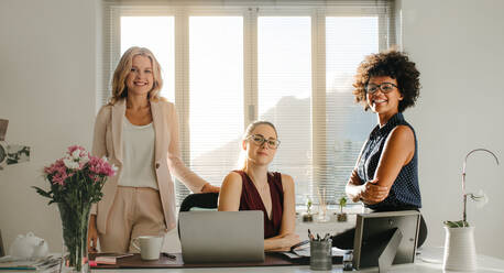 Drei Geschäftsfrauen in Freizeitkleidung am Schreibtisch, die in die Kamera schauen und lächeln. Erfolgreiche Startup-Geschäftspartner im Büro. - JLPSF16913