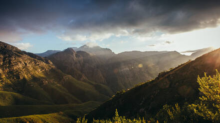 Sonnenstrahlen von rechts über dem Hügel, die das Bergtal erhellen. Berge und bewölkter Himmel bei Sonnenuntergang im Naturschutzgebiet Jonkershoek. - JLPSF16962