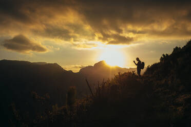 Silhouette eines Mannes, der mit seinem Smartphone ein Bergtal bei Sonnenuntergang fotografiert. Wanderer, der im Naturschutzgebiet Jonkershoek fotografiert, während im Hintergrund die Sonne über den Bergen scheint. - JLPSF16965