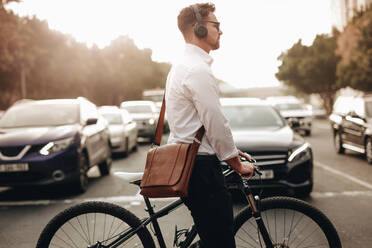 Businessman walking on road holding his bicycle crossing the road. Man wearing office bag and wireless headphones going to office holding his bike. - JLPSF16969