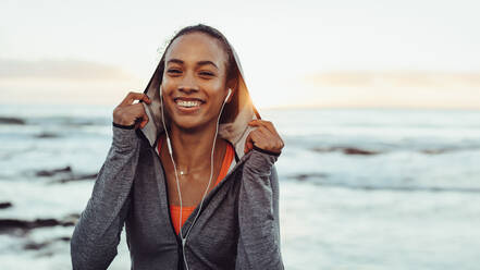 Positive young woman runner in sportswear and earphones outdoors against sea. Female resting after workout by the sea. - JLPSF16973