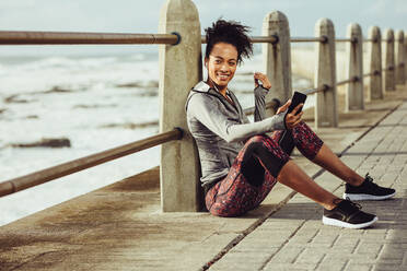 Happy young fit woman sitting at seaside promenade and listening to music from her mobile phone. Female runner resting after her training session. - JLPSF16987