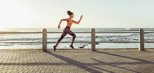 Sportliche junge Frau beim Laufen auf der Strandpromenade, Seitenansicht einer Läuferin beim Sprint im Freien. - JLPSF16988