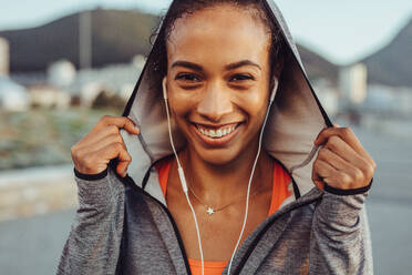 Nahaufnahme einer lächelnden jungen Frau in Sportkleidung und mit Kopfhörern. Selbstbewusste Fitness-Frau in einem Hoodie. - JLPSF16990