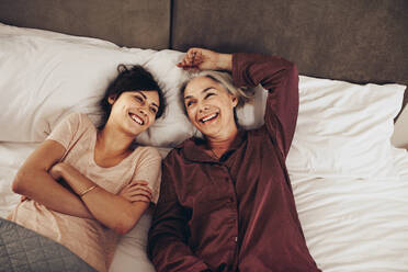 Lächelnde Frau, die neben ihrer alten Mutter mit verschränkten Armen auf dem Bett schläft. Mutter und Tochter verbringen glückliche Zeit miteinander, während sie auf dem Bett zu Hause liegen. - JLPSF16992