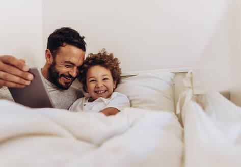Mann liegt mit seinem Sohn auf dem Bett und unterhält sich. Fröhlicher Vater und Sohn verbringen Zeit miteinander und benutzen einen Tablet-PC. - JLPSF16998