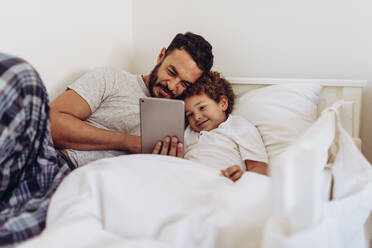 Vater und Sohn liegen auf dem Bett und schauen sich einen Film auf einem Tablet-PC an. Ein Mann liegt auf dem Bett und sein Kind hält einen Tablet-PC in der Hand. - JLPSF16999