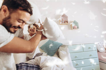 Cheerful man pillow fighting with son sitting on bed at home. Father and son having fun playing with pillows with feathers flying around. - JLPSF17001