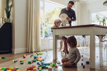 Ein Mann, der sich um seine Kinder kümmert, während er den Haushalt erledigt. Ein Kind, das mit Spielzeug spielt und auf dem Boden sitzt, während sein Vater die tägliche Hausarbeit erledigt. - JLPSF17006