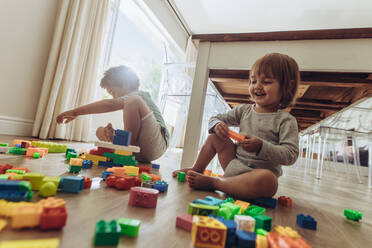 Glückliche Kinder spielen mit Bauklötzen auf dem Boden sitzend. Zwei Kinder spielen mit Spielzeug zu Hause. - JLPSF17009