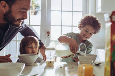 Ein Mann beobachtet sein Kind, wie es Milch in seine Frühstücksschüssel gießt. Vater und Kinder sitzen am Tisch und bereiten das Frühstück vor. - JLPSF17014