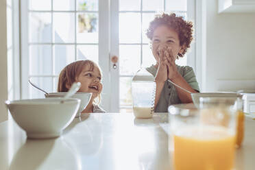 Zwei Kinder sitzen am Frühstückstisch mit Milchflasche und Saft auf dem Tisch. Fröhliche Kinder haben Spaß und lächeln am Frühstückstisch. - JLPSF17015