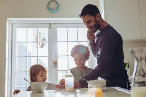 Ein Mann steht am Frühstückstisch und spricht mit seinen Kindern über das Handy. Ein Mann passt auf seine Kinder auf und verwaltet die Büroarbeit von zu Hause aus über das Handy. - JLPSF17016