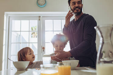 Ein Vater, der zu Hause seine Kinder beaufsichtigt und mit dem Handy Büroarbeit erledigt. Ein Mann, der mit seinen Kindern am Frühstückstisch steht. - JLPSF17017
