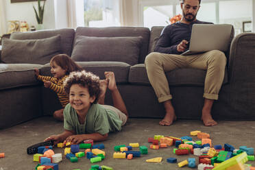 Ein Mann arbeitet am Laptop, während seine Kinder auf dem Boden spielen. Fröhliche Kinder spielen mit Bauklötzen und sehen zu Hause fern. - JLPSF17023