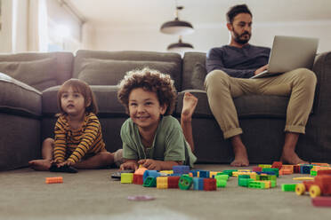 Lächelnde Kinder, die zu Hause mit Bauklötzen spielen und fernsehen. Ein Mann, der auf seine Kinder aufpasst und von zu Hause aus arbeitet. - JLPSF17024